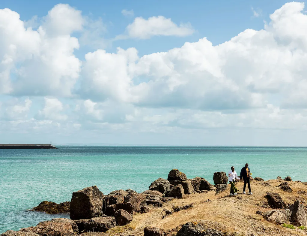 Two people walking along a rocky coastline beside turquoise sea under large white clouds.