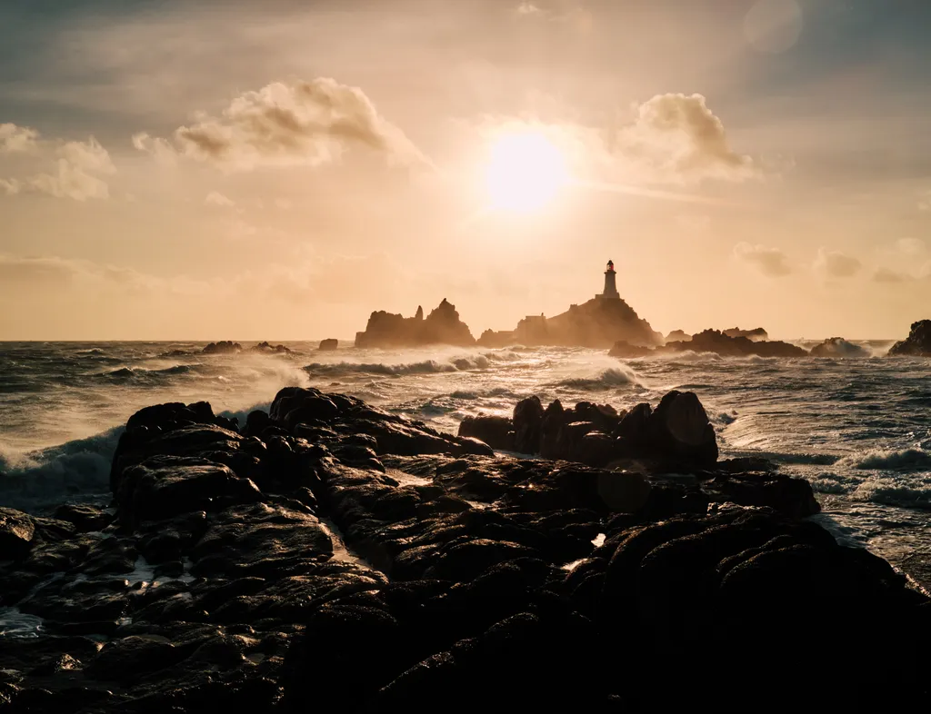 Corbière Lighthouse silhouetted against a glowing sunset above a rugged sea and dark rocky shoreline in Jersey