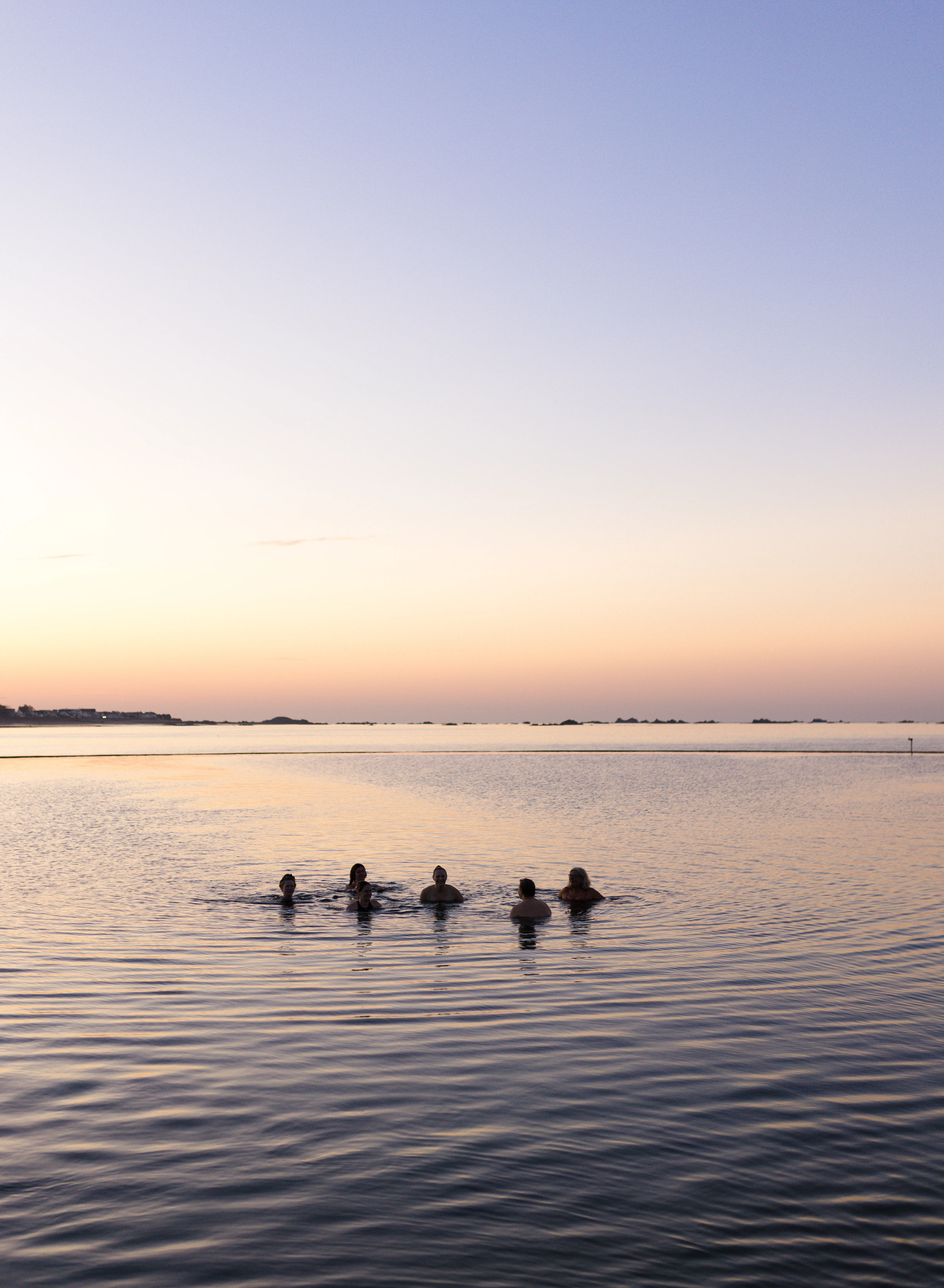 Six people swimming in calm sea water at sunset beneath a pastel sky.