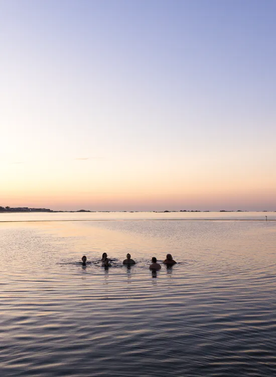 Six people swimming in calm sea water at sunset beneath a pastel sky.
