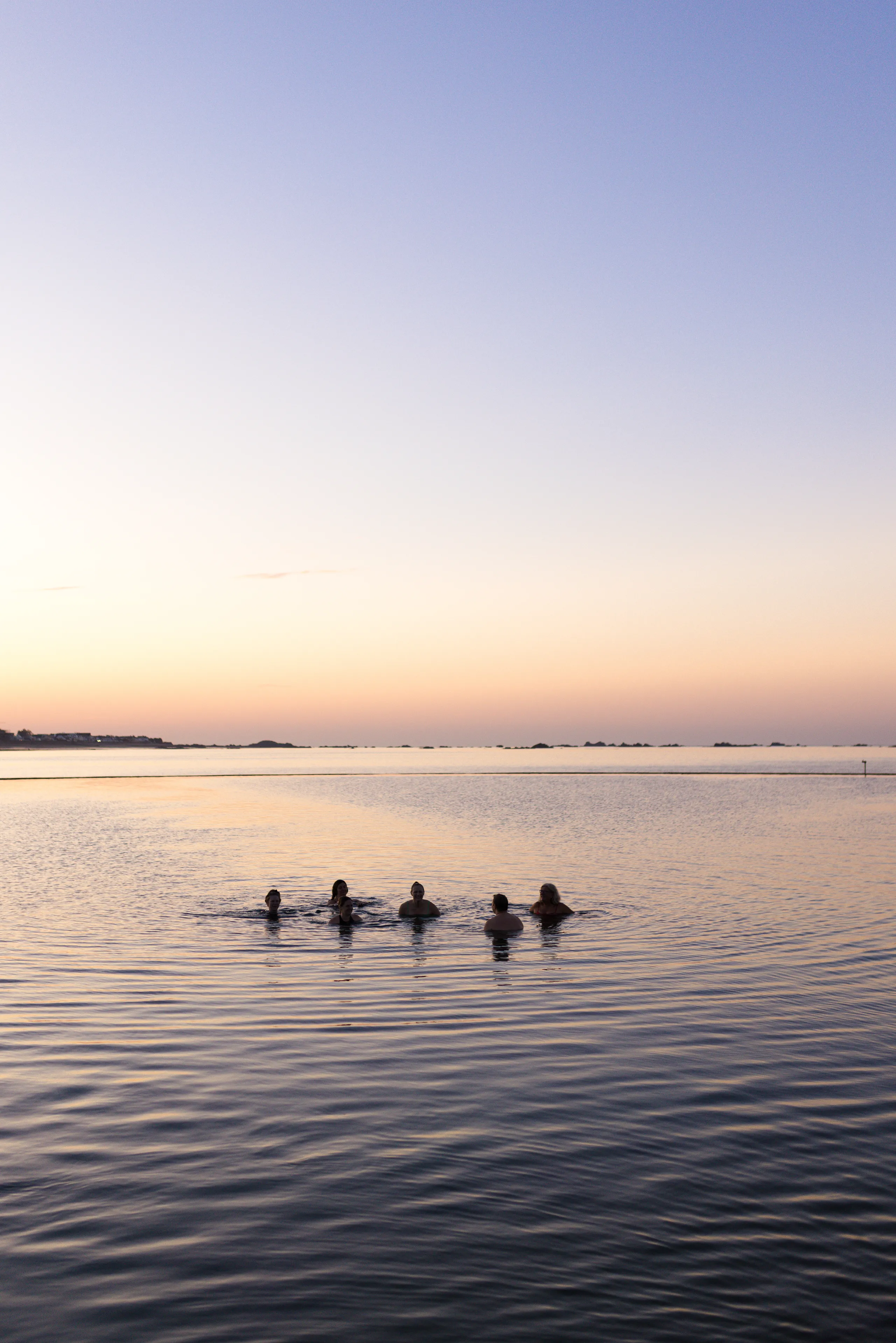 Six people swimming in calm sea water at sunset beneath a pastel sky.