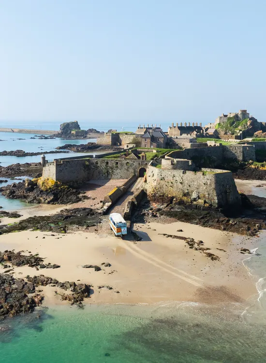 Aerial view of a historic island fortress connected to a sandy beach by a stone causeway, surrounded by clear turquoise sea and rocky shoreline.