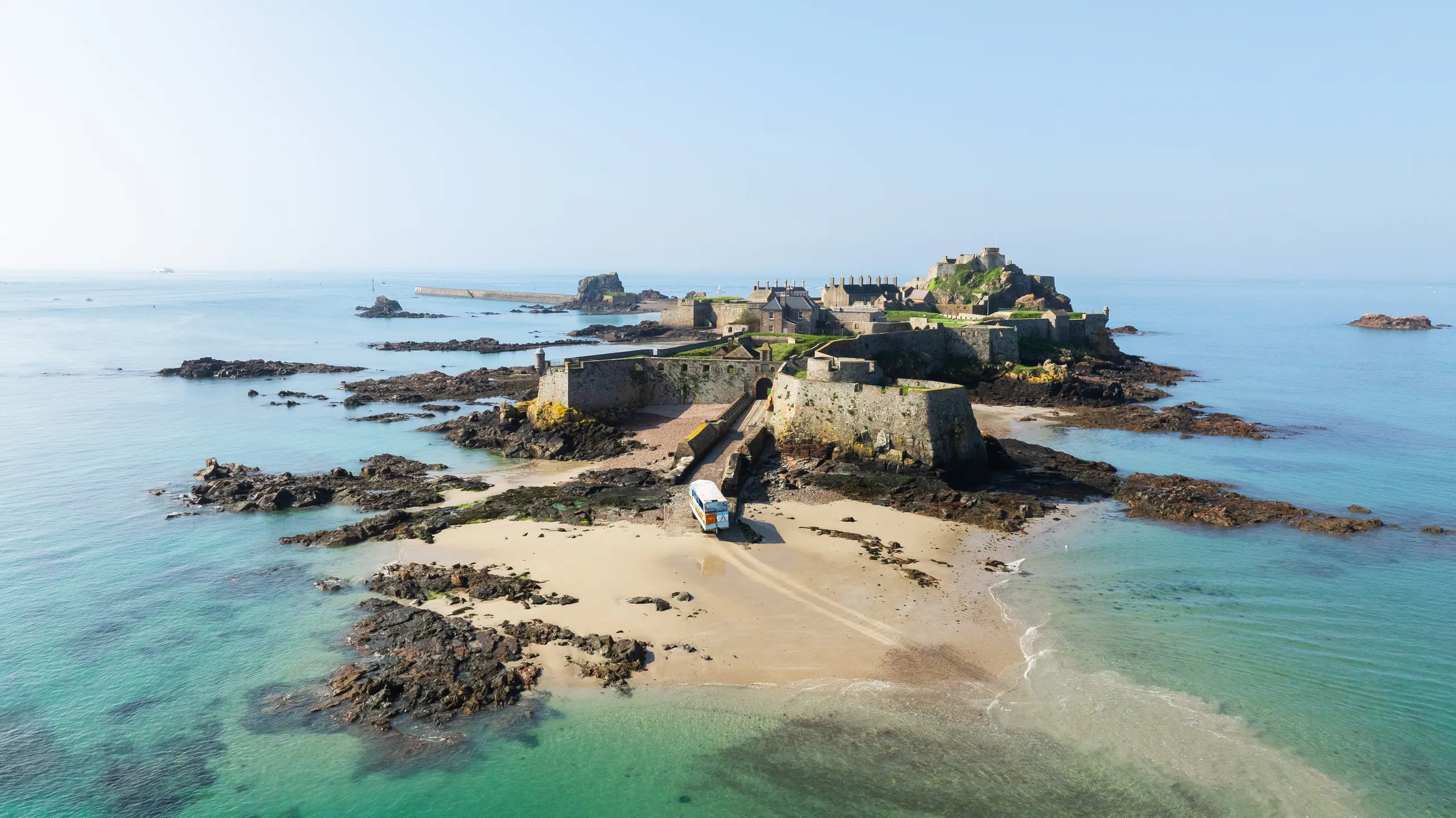 Aerial view of a historic island fortress connected to a sandy beach by a stone causeway, surrounded by clear turquoise sea and rocky shoreline.