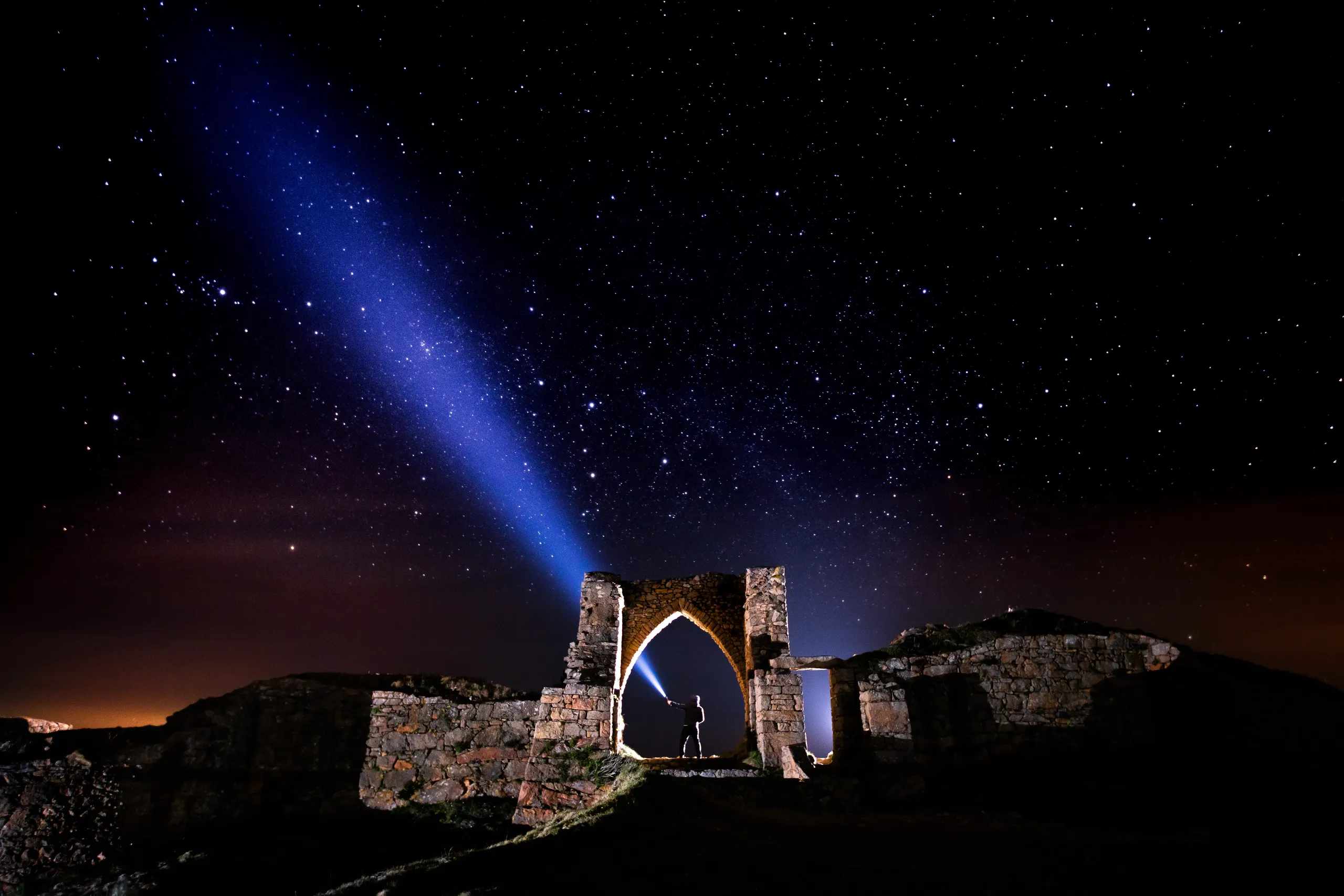 Silhouette of a person shining a torch through a stone archway beneath a star-filled night sky