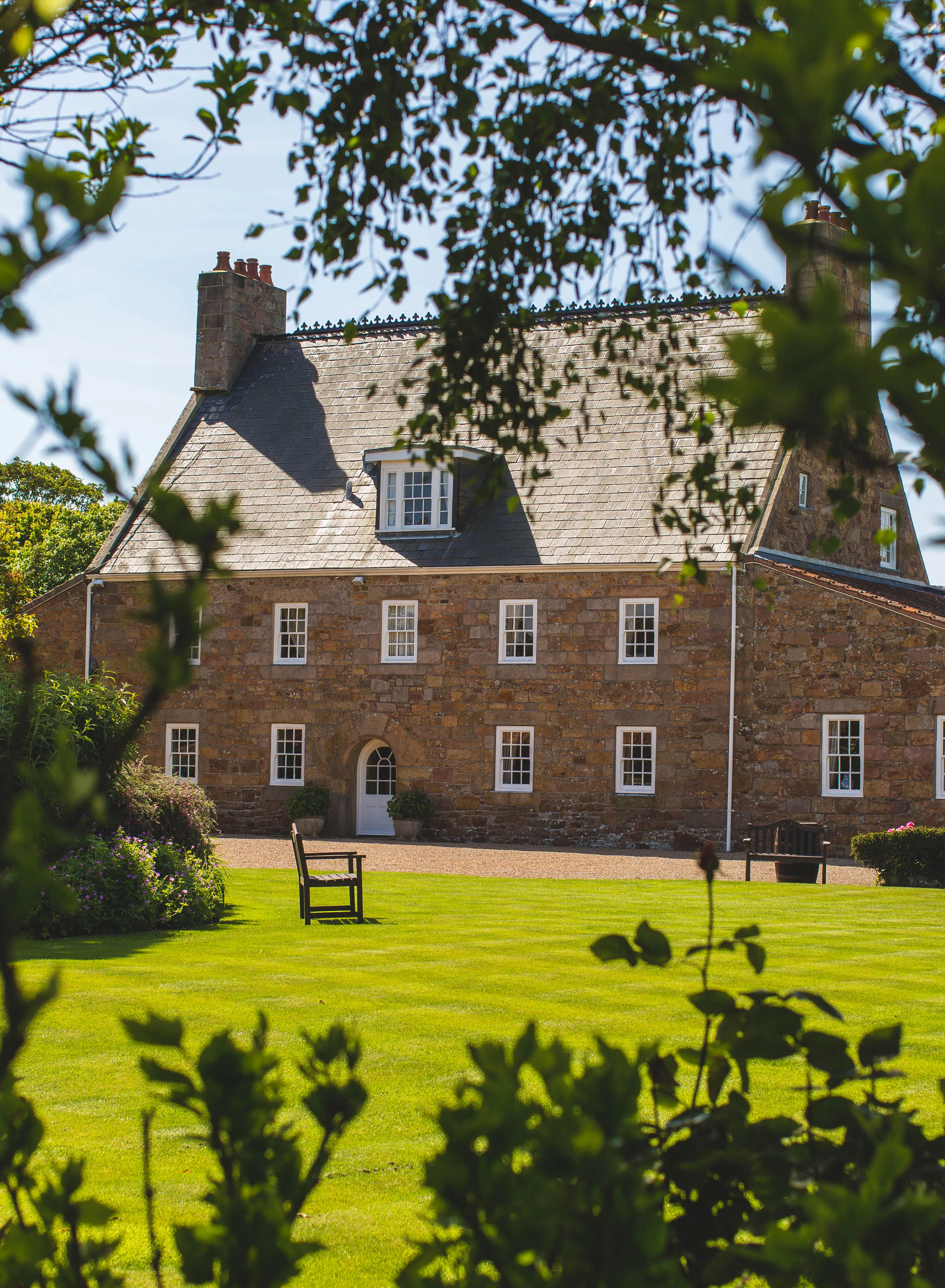 Traditional stone house viewed through leafy garden branches on a sunny day