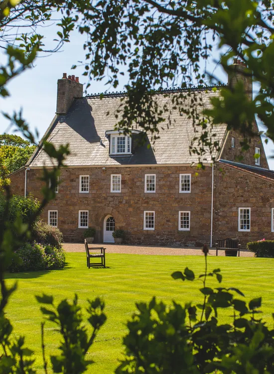 Traditional stone house viewed through leafy garden branches on a sunny day