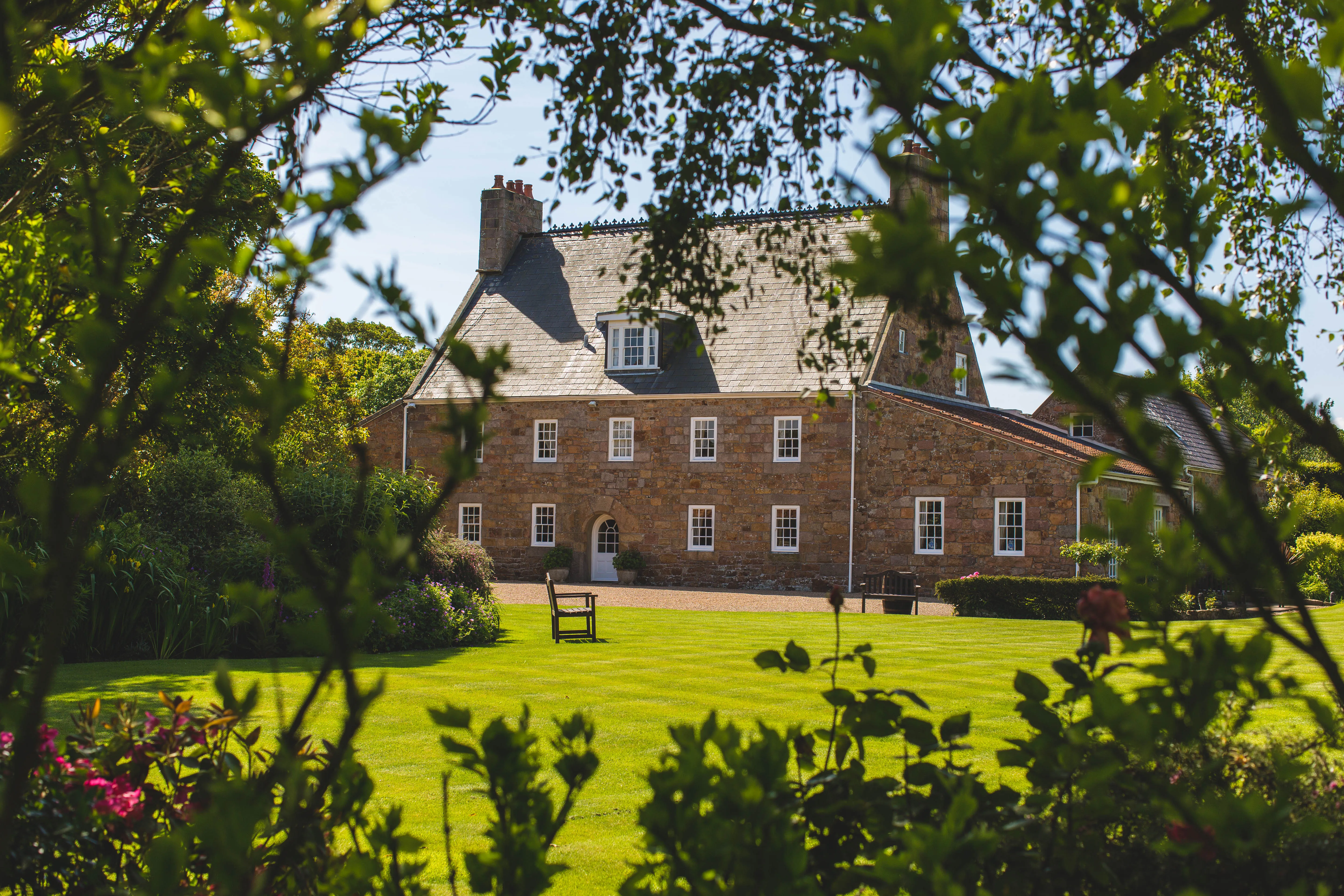 Traditional stone house viewed through leafy garden branches on a sunny day