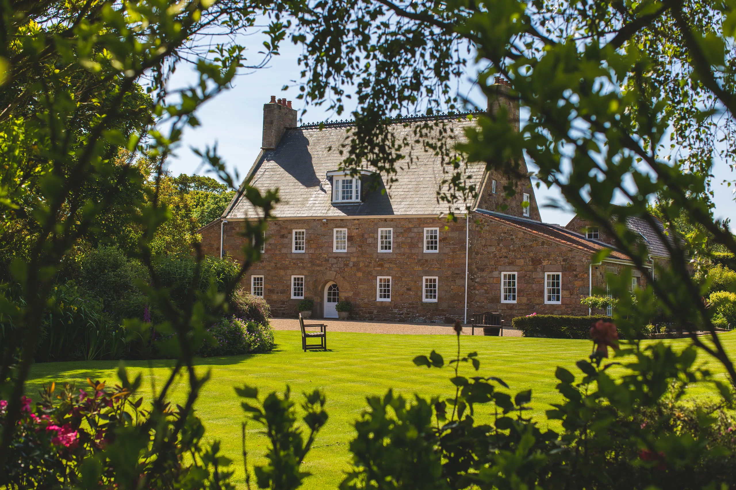 Traditional stone house viewed through leafy garden branches on a sunny day