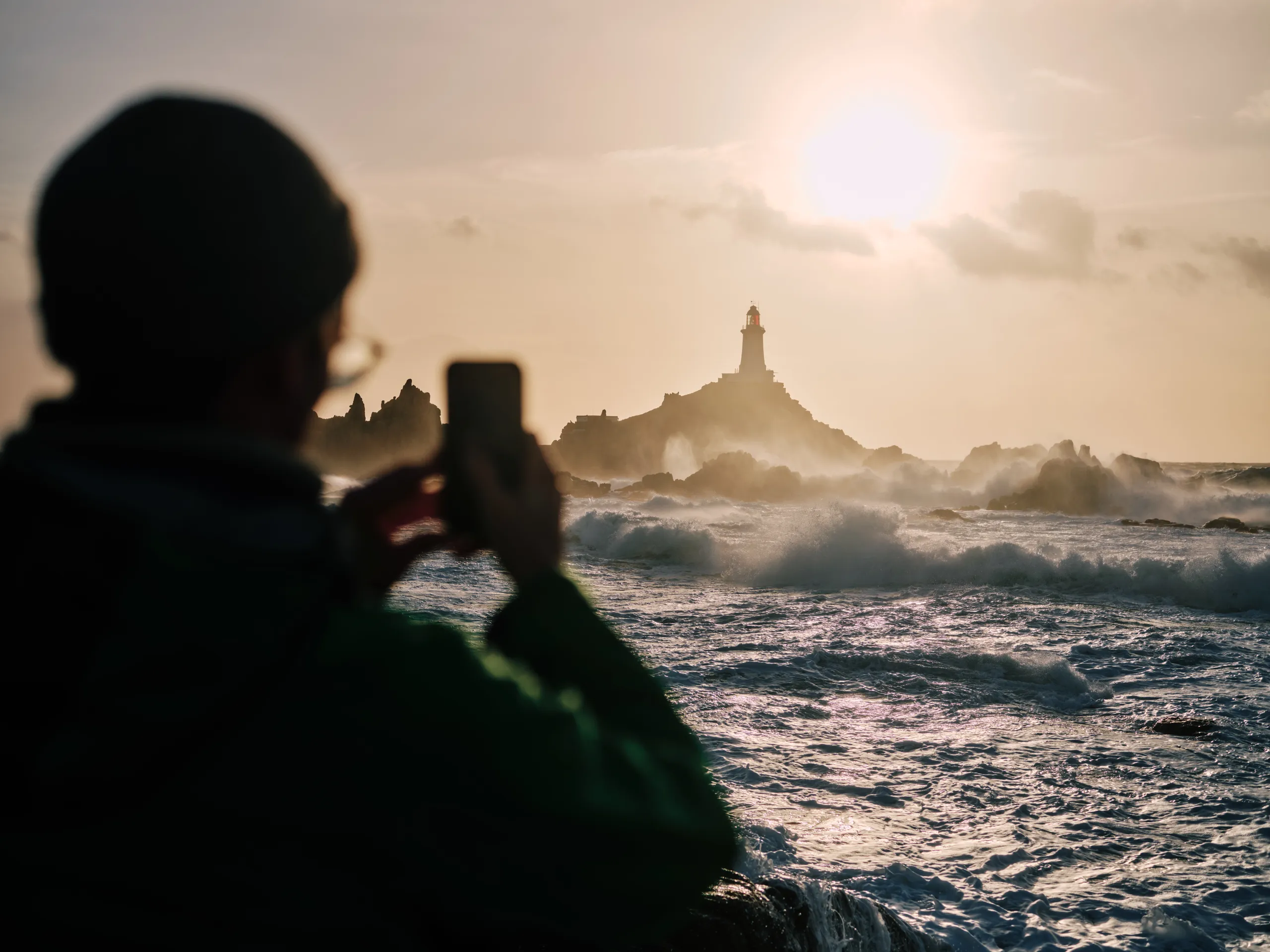 Silhouetted visitor photographing Corbière Lighthouse as waves crash against the rocky Jersey coastline at sunset.