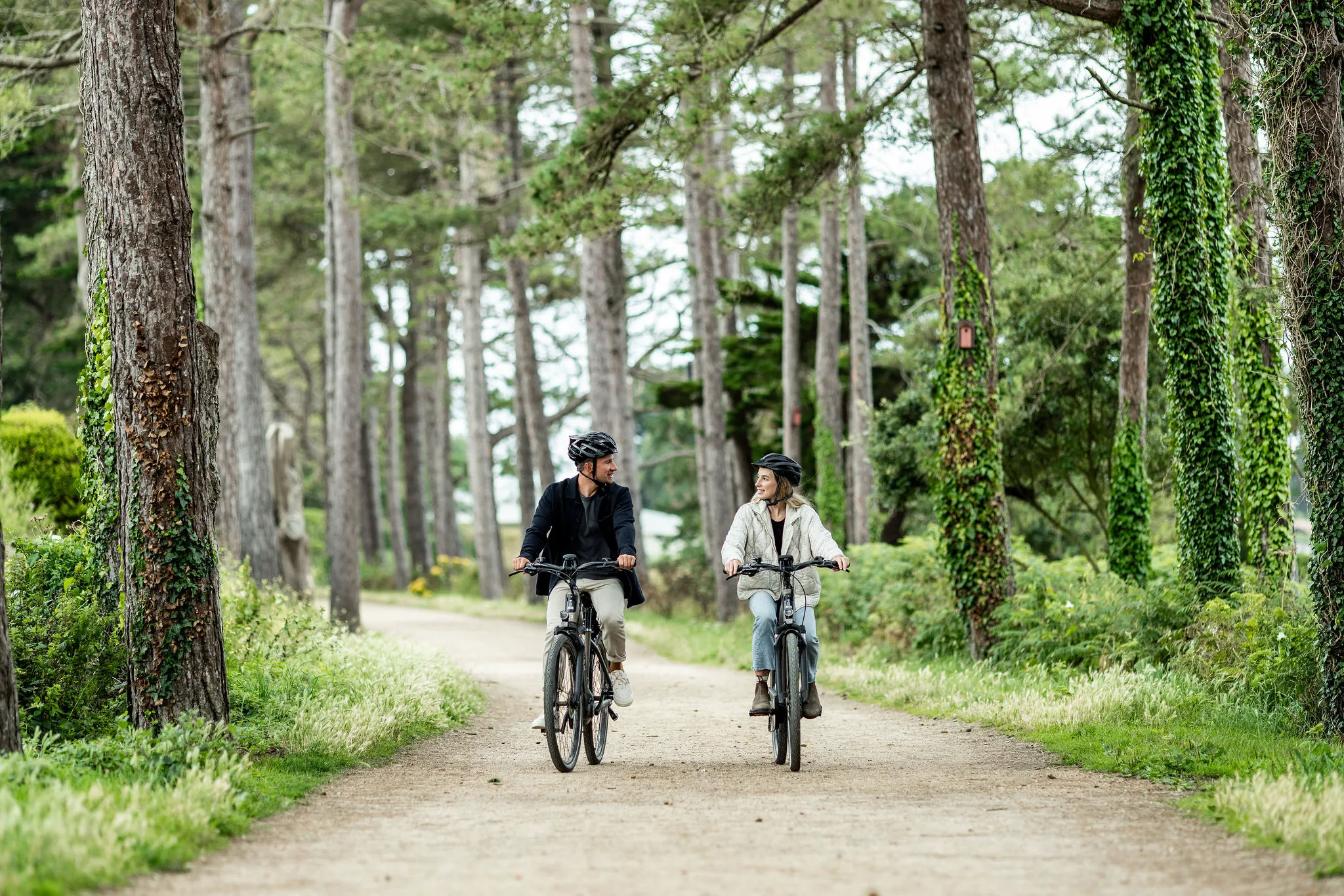 Two cyclists riding side by side along a tree-lined gravel path in a lush woodland setting.