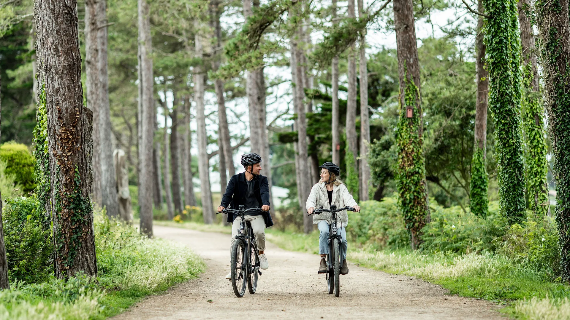 Two cyclists riding side by side along a tree-lined gravel path in a lush woodland setting.
