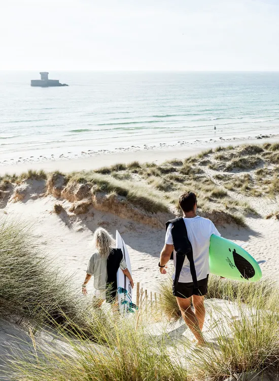 Two surfers walk through sandy dunes towards St Ouen's Bay beach with surfboards on a bright day.