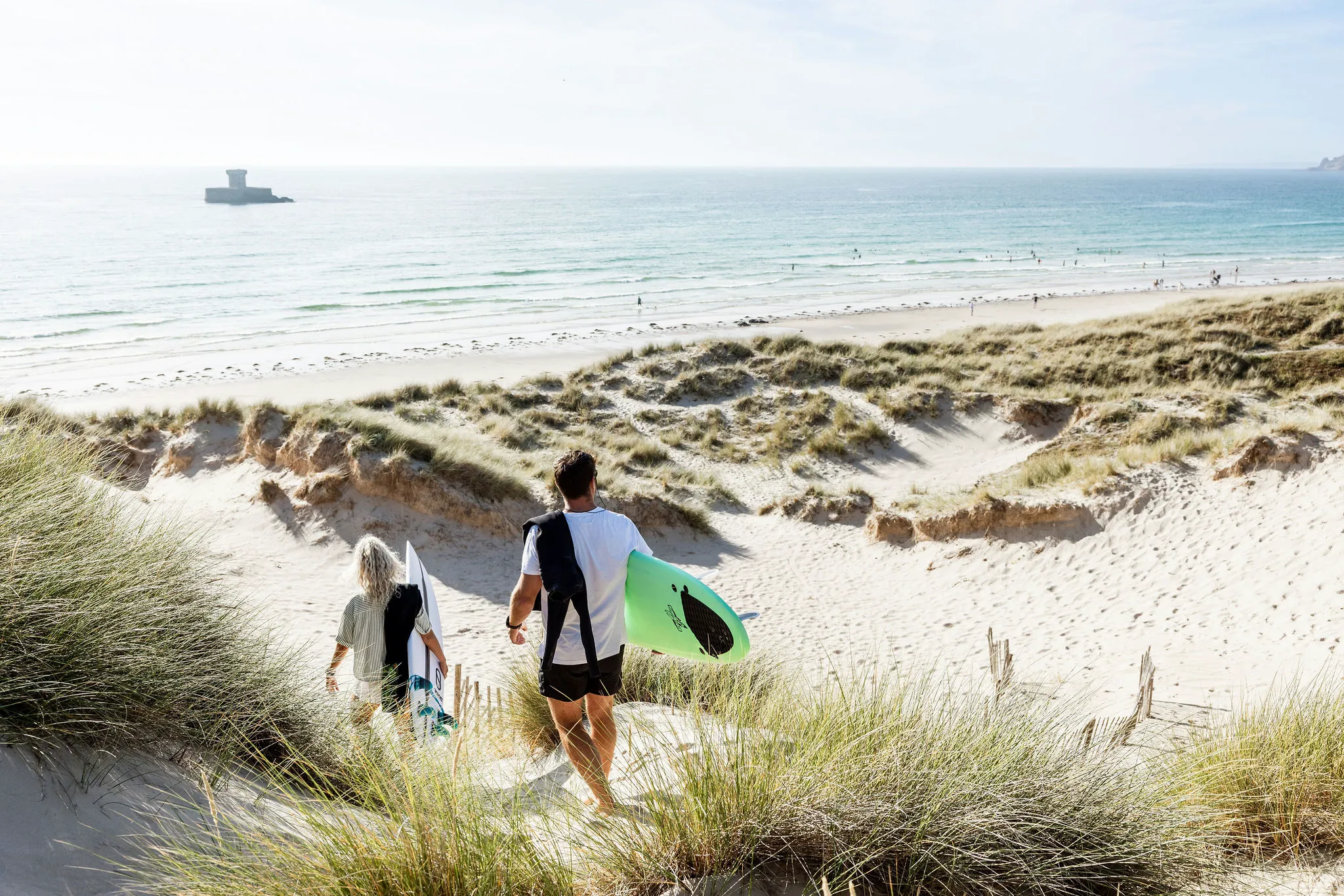 Two surfers walk through sandy dunes towards St Ouen's Bay beach with surfboards on a bright day.
