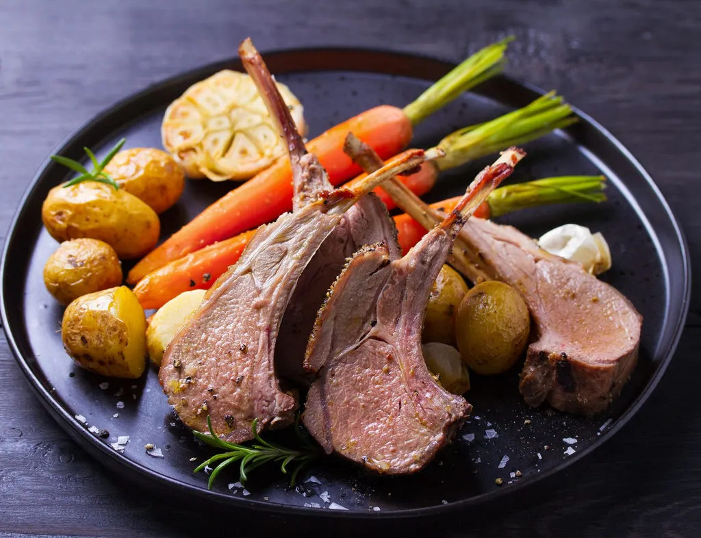 Roasted rack of lamb served with baby potatoes, carrots, roasted garlic and rosemary on a black plate.