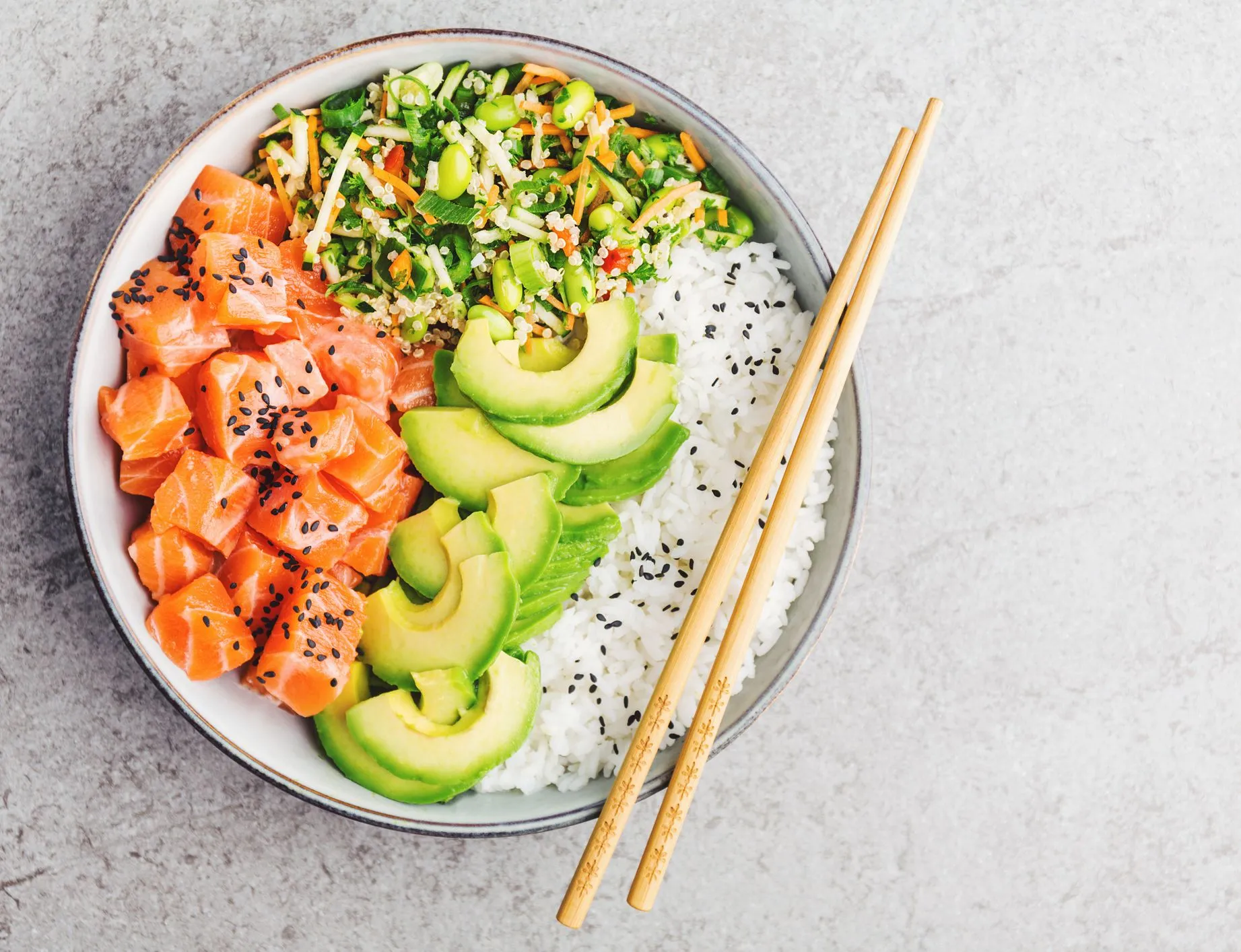 Overhead view of a salmon poke bowl with rice, avocado, edamame, mixed vegetables and black sesame seeds, with chopsticks on the side