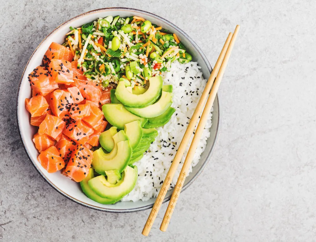 Overhead view of a salmon poke bowl with rice, avocado, edamame, mixed vegetables and black sesame seeds, with chopsticks on the side