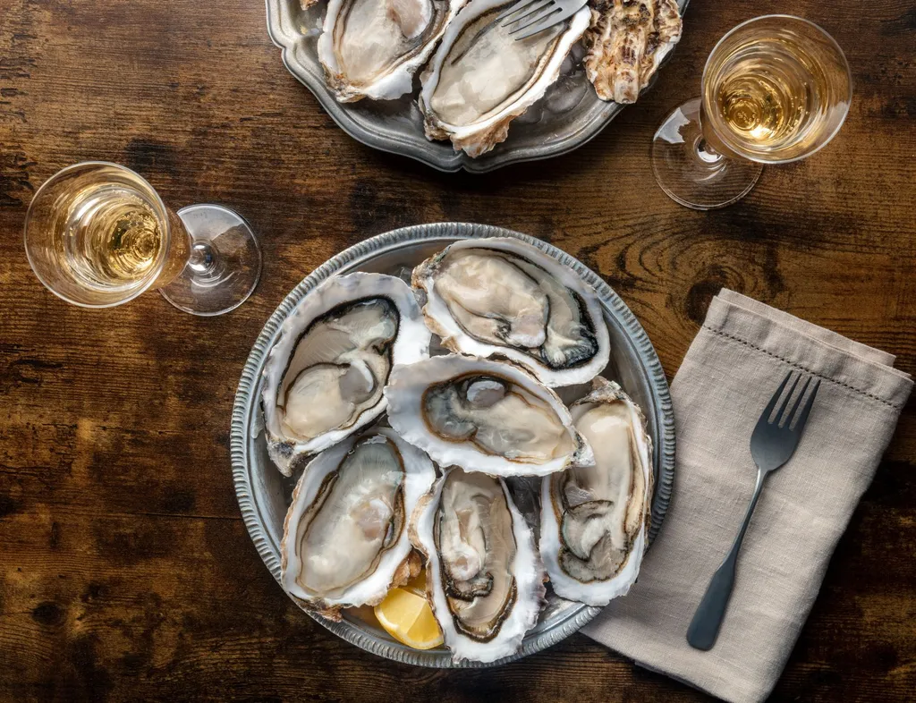 Overhead view of fresh oysters on silver platters with lemon wedges and two glasses of white wine on a wooden table