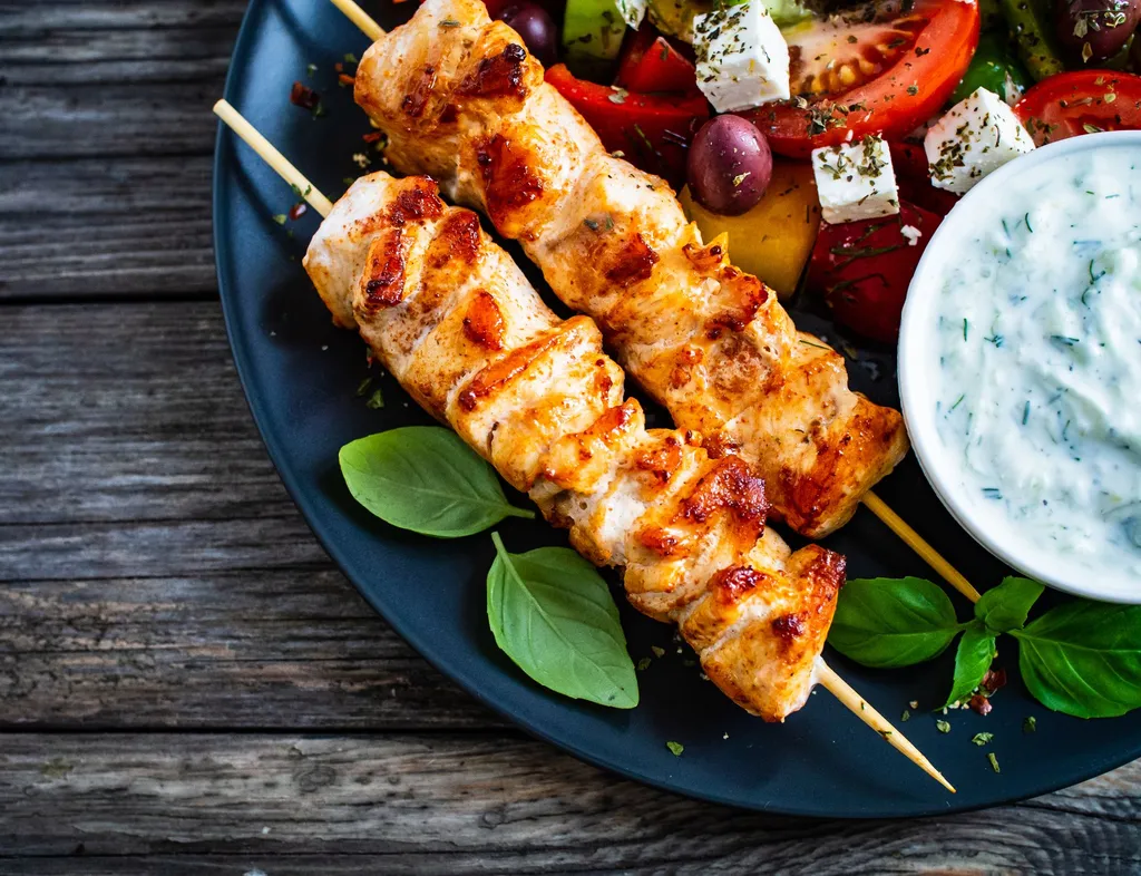 Grilled chicken skewers served with a Greek-style salad, basil leaves and a bowl of creamy yoghurt dip on a dark plate.