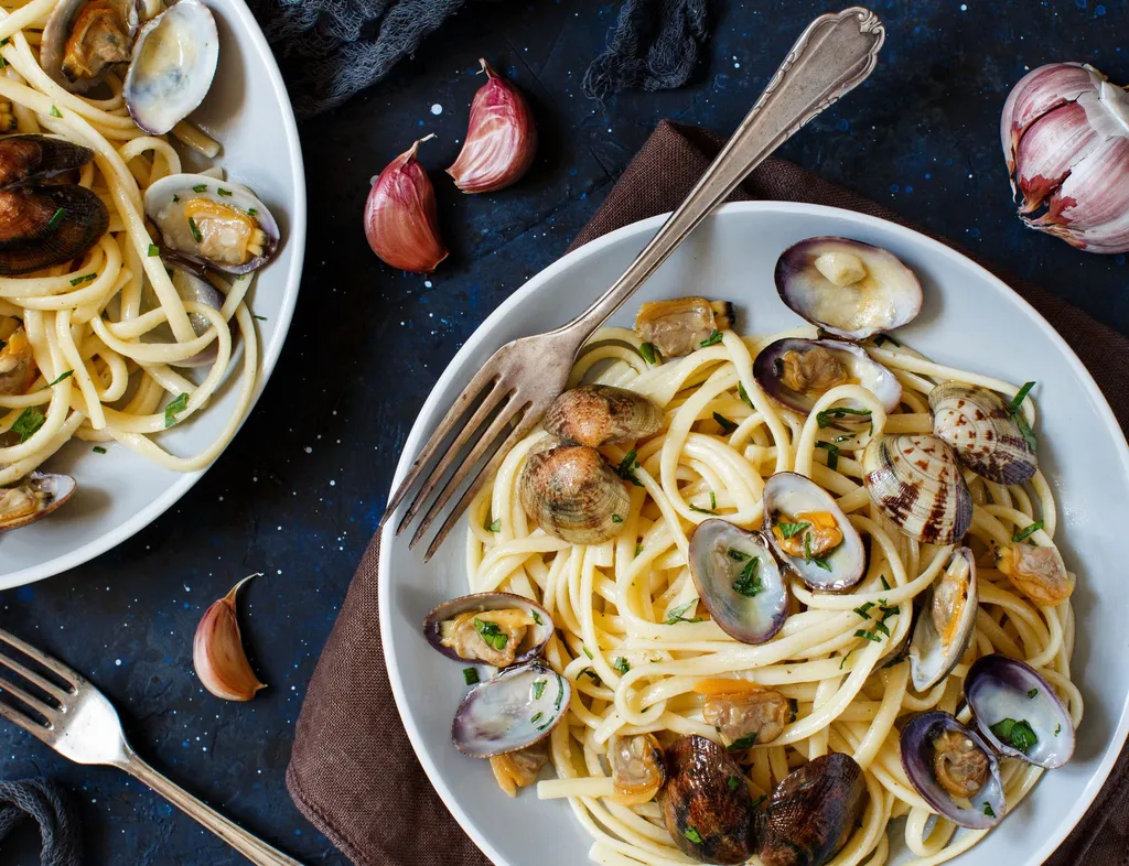Two plates of spaghetti with clams, served with garlic and forks on a dark table.