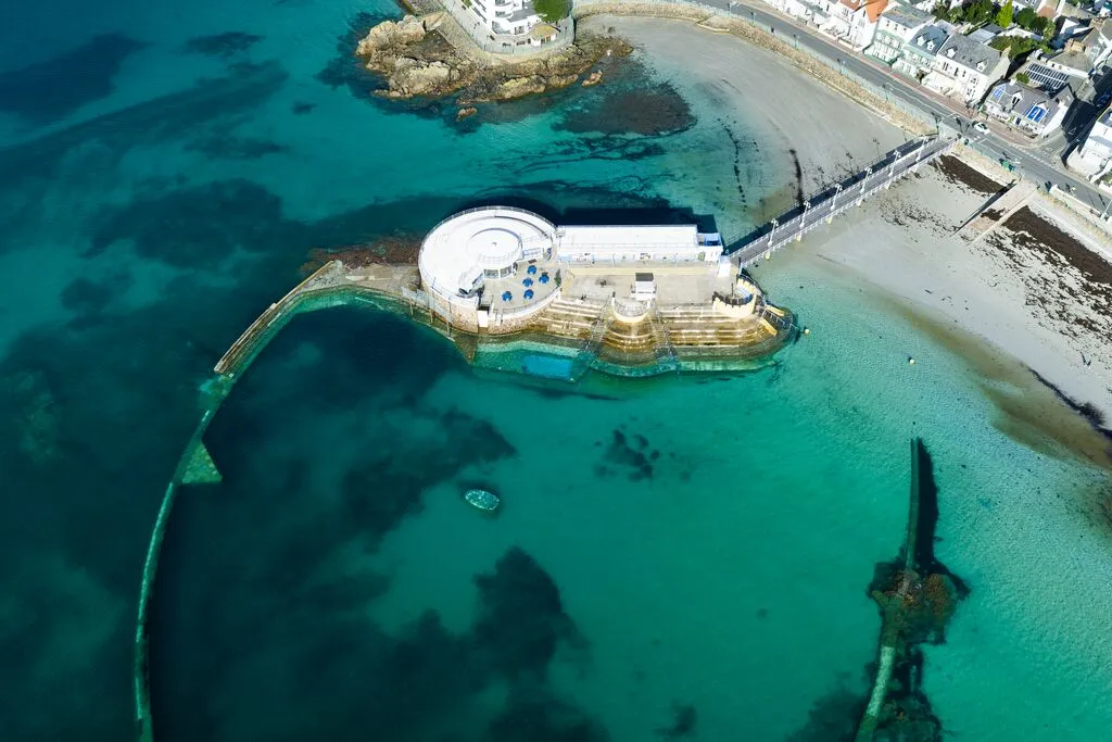 Aerial view of a seaside tidal pool complex with curved sea walls, clear turquoise water and a sandy beach beside the promenade.