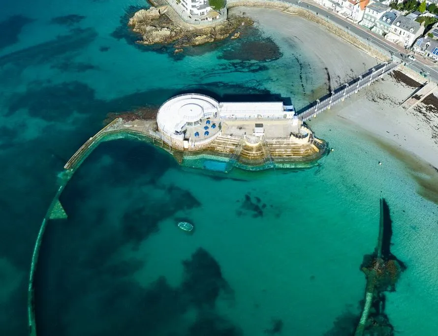 Aerial view of a seaside tidal pool complex with curved sea walls, clear turquoise water and a sandy beach beside the promenade.