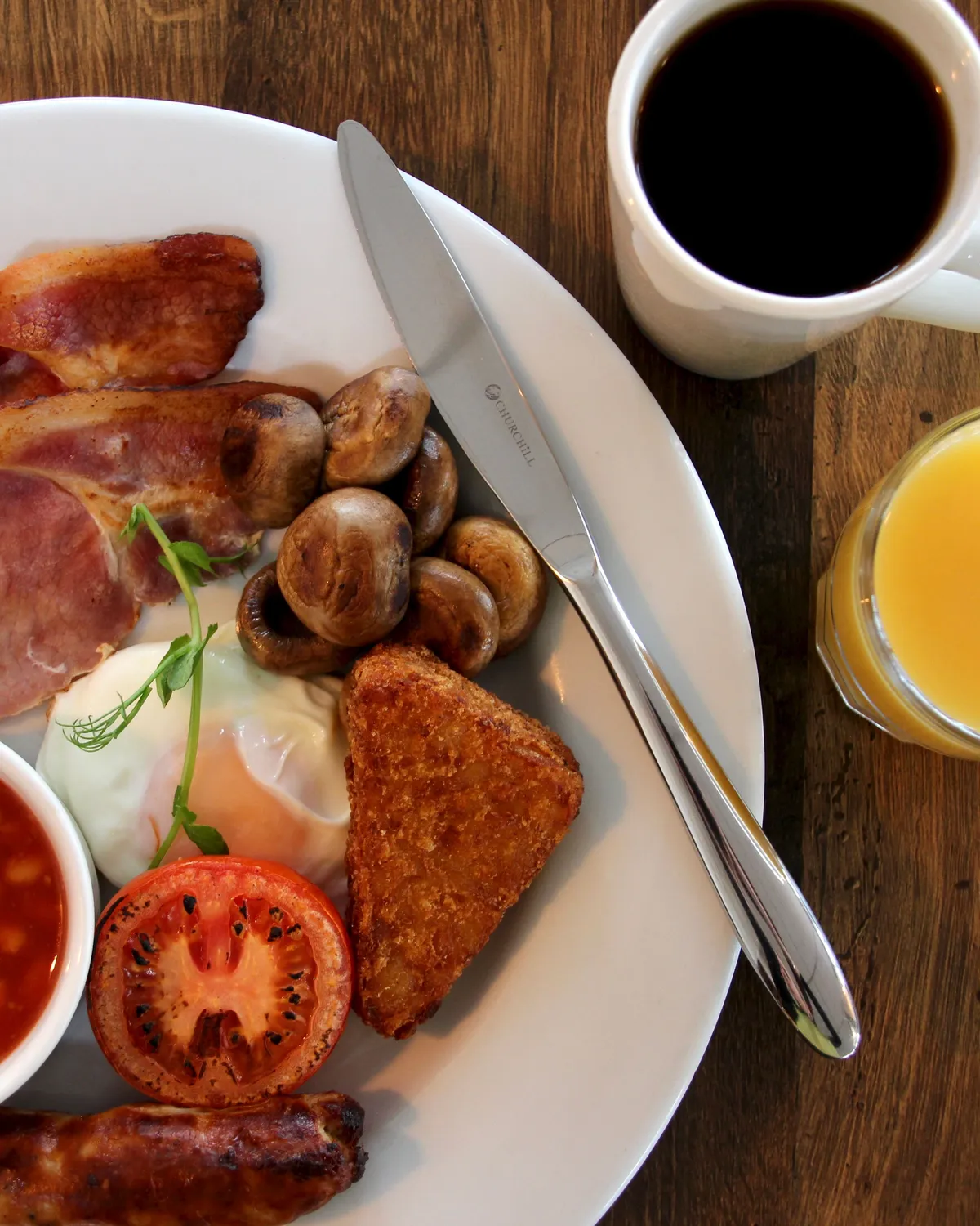 Cooked breakfast with bacon, mushrooms, tomato, beans, egg, hash brown and sausage, served with black coffee and orange juice on a wooden table
