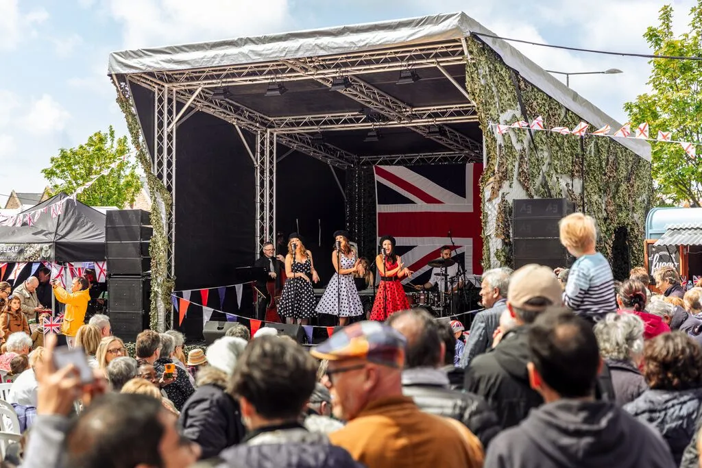 Outdoor live music performance on a covered stage with a large crowd and Union Jack backdrop