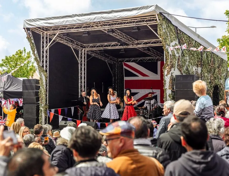 Outdoor live music performance on a covered stage with a large crowd and Union Jack backdrop