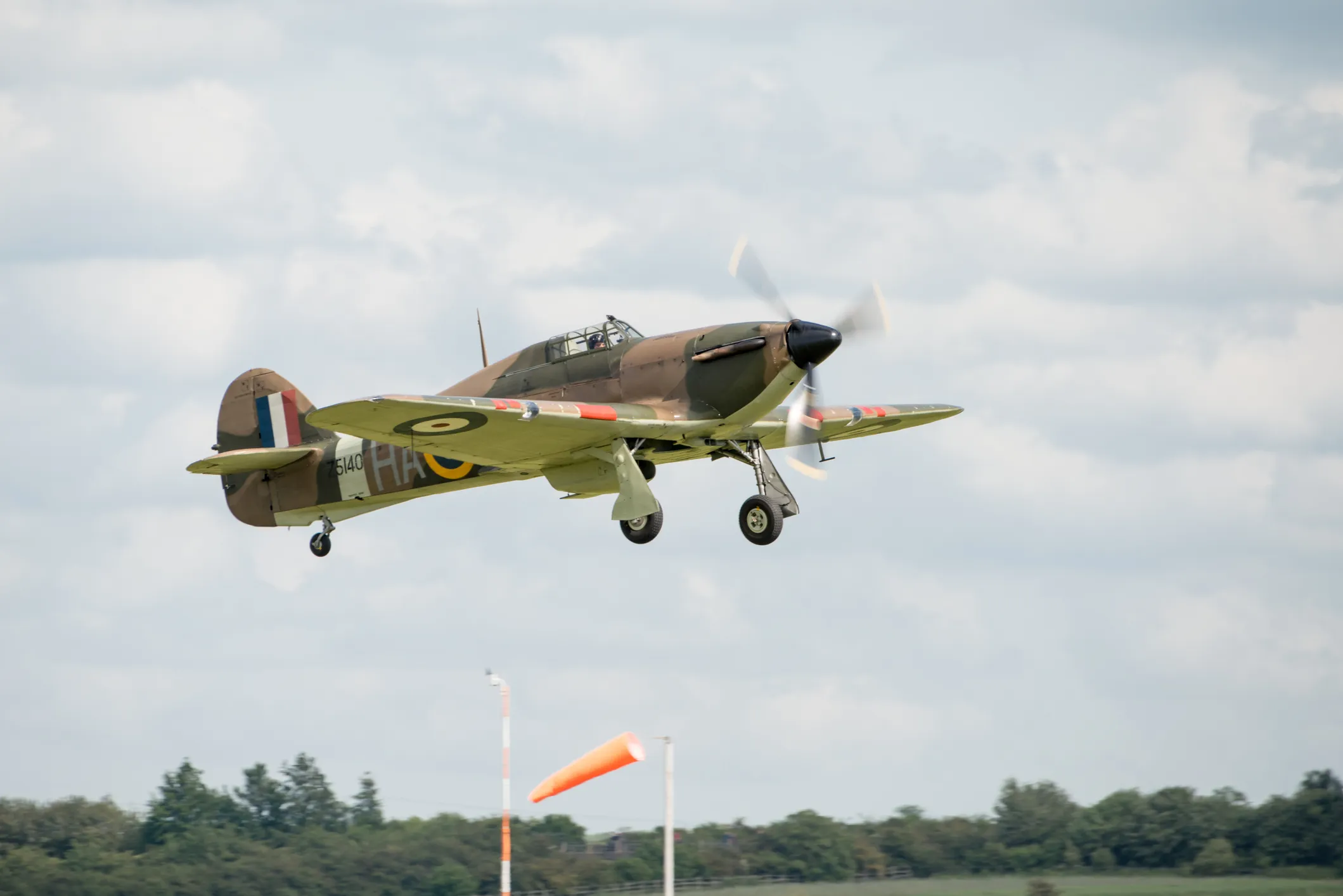 Vintage RAF fighter aircraft in camouflage livery flying with landing gear down against a cloudy sky