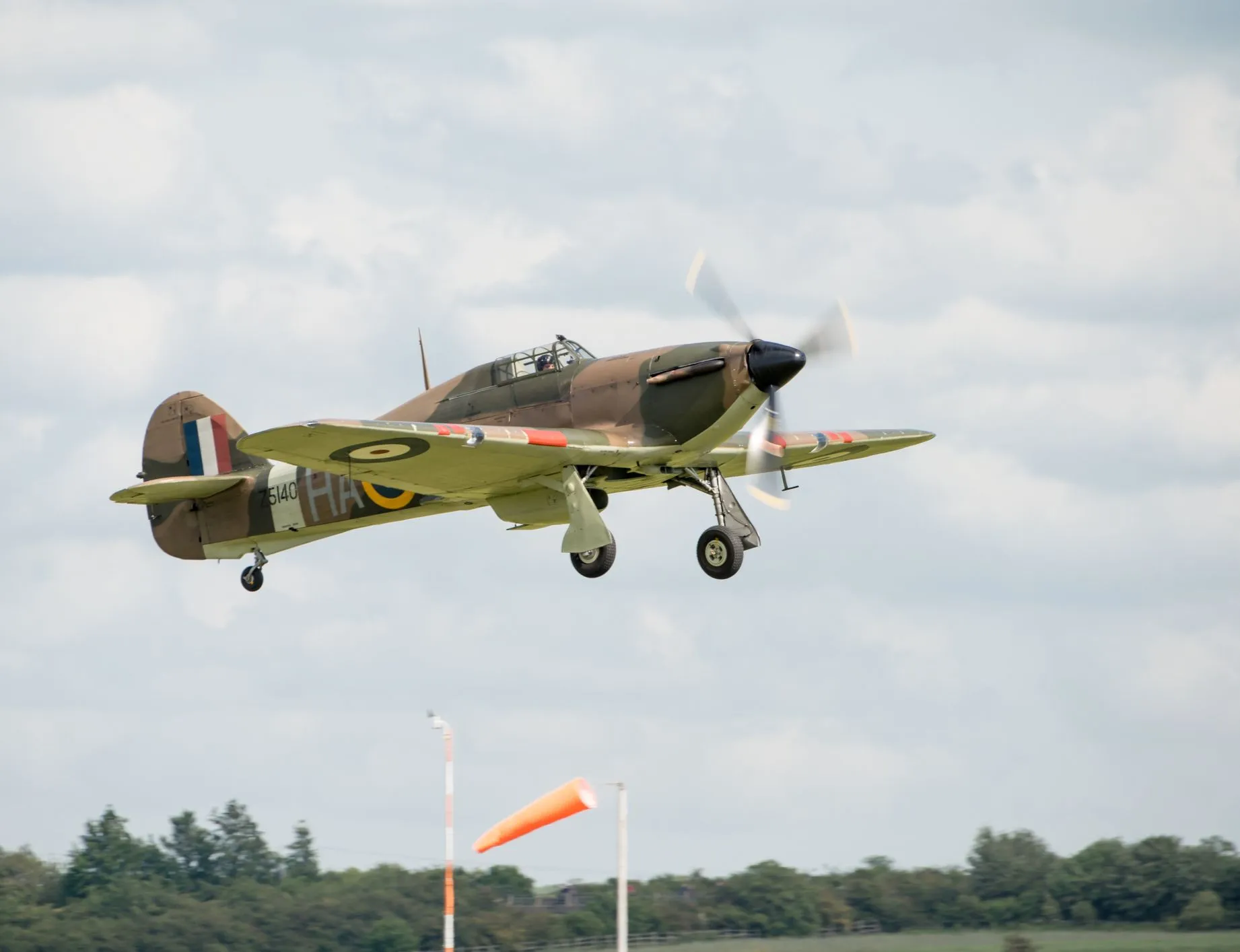 Vintage RAF fighter aircraft in camouflage livery flying with landing gear down against a cloudy sky