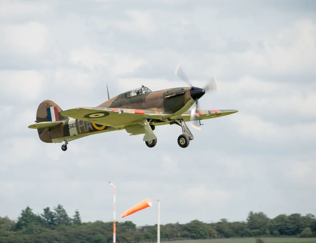 Vintage RAF fighter aircraft in camouflage livery flying with landing gear down against a cloudy sky