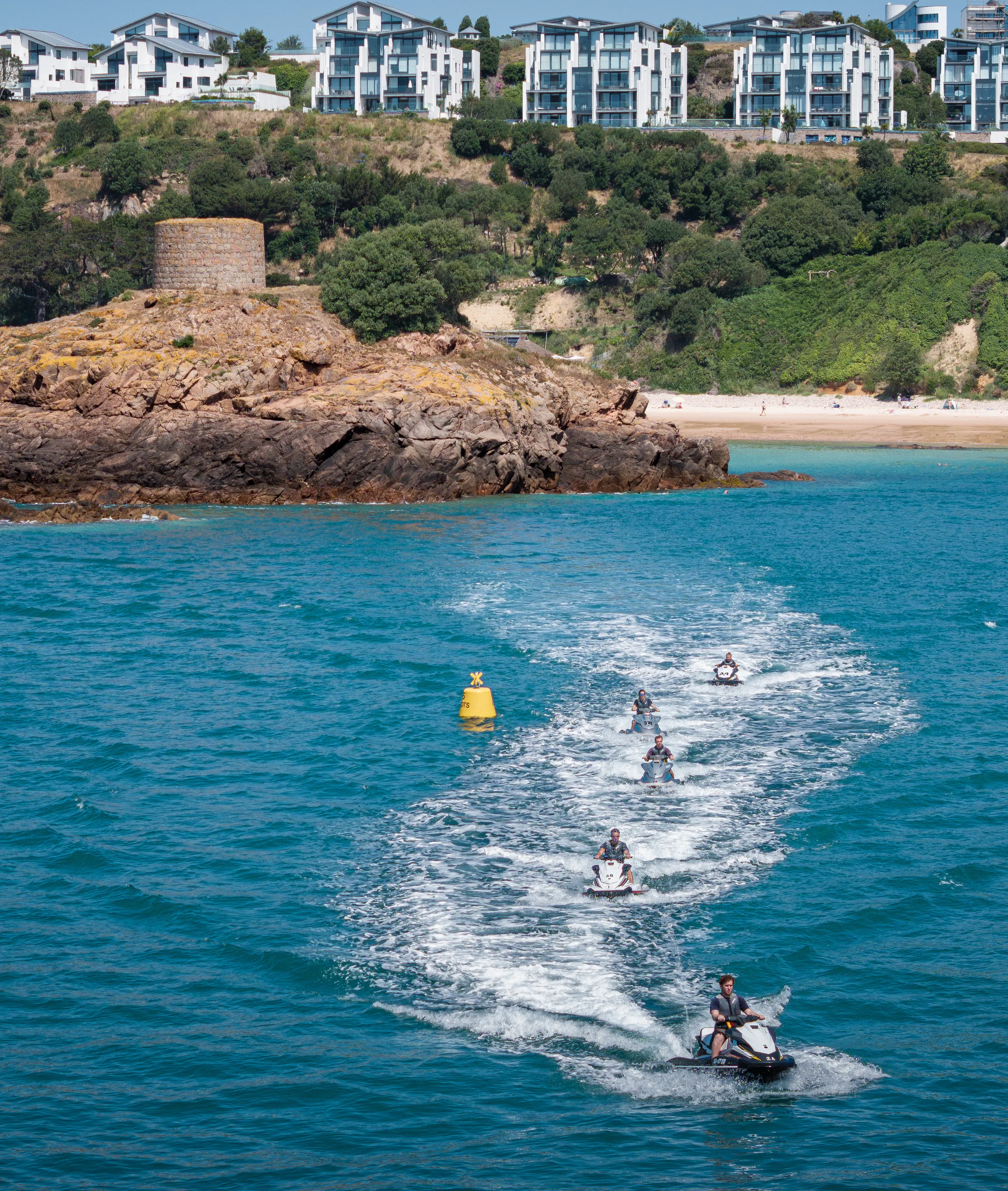 A line of jet skis speeds across bright blue coastal water towards a rocky headland with a stone tower, beach and hillside apartments behind.