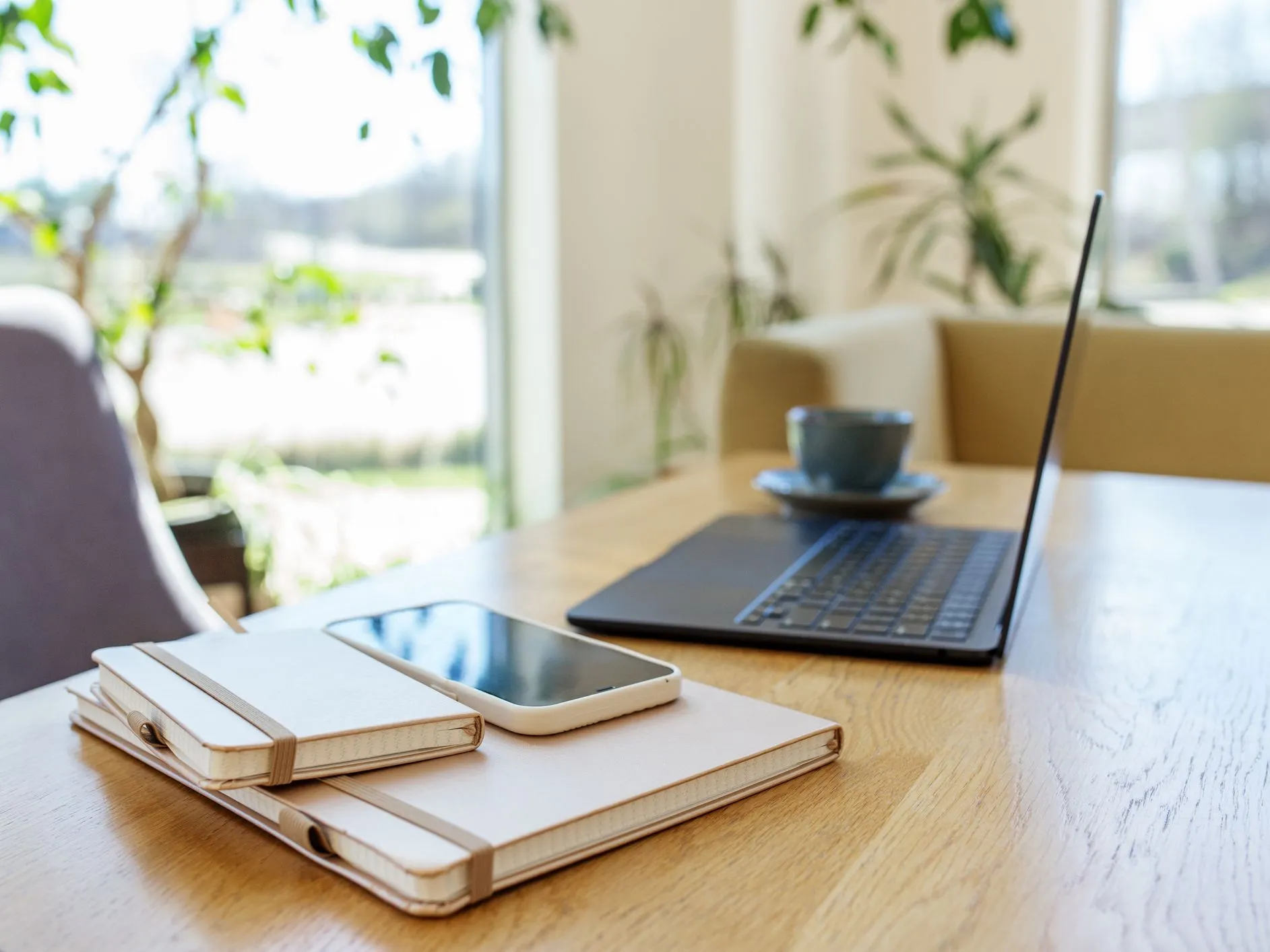 Laptop, notebooks, smartphone and a cup on a wooden table in a bright home office