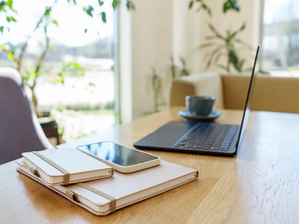 Laptop, notebooks, smartphone and a cup on a wooden table in a bright home office