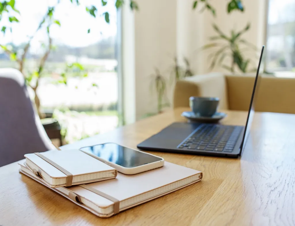 Laptop, notebooks, smartphone and a cup on a wooden table in a bright home office