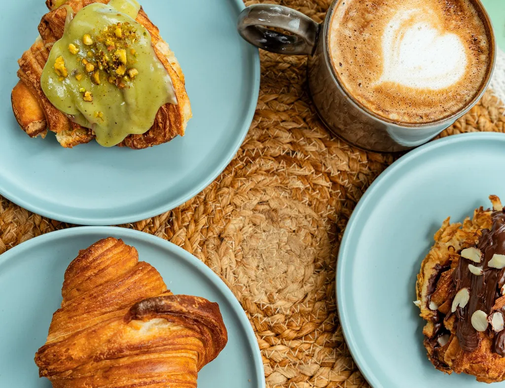 Overhead view of croissants on pale blue plates with pistachio icing, chocolate topping and a frothy coffee on a woven placemat.