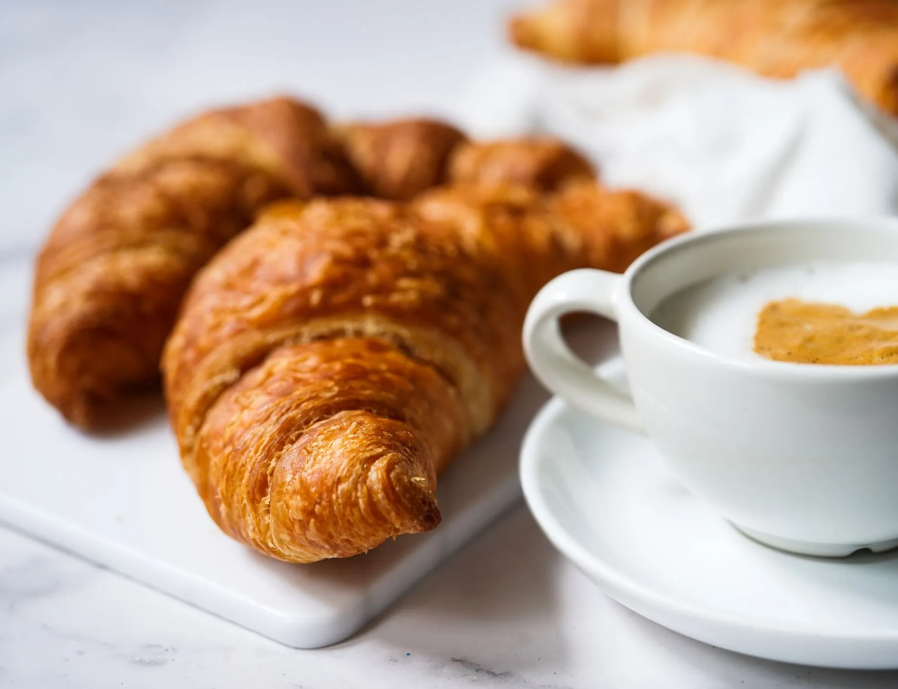 Golden croissants on a white board beside a cup of coffee with milk foam
