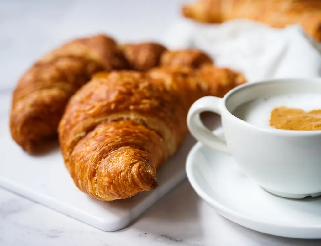 Golden croissants on a white board beside a cup of coffee with milk foam