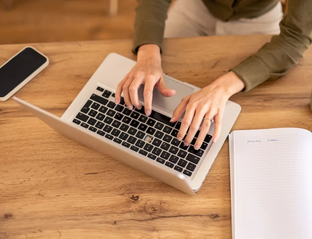 Person typing on a laptop at a wooden desk with a smartphone, notebook and mug nearby