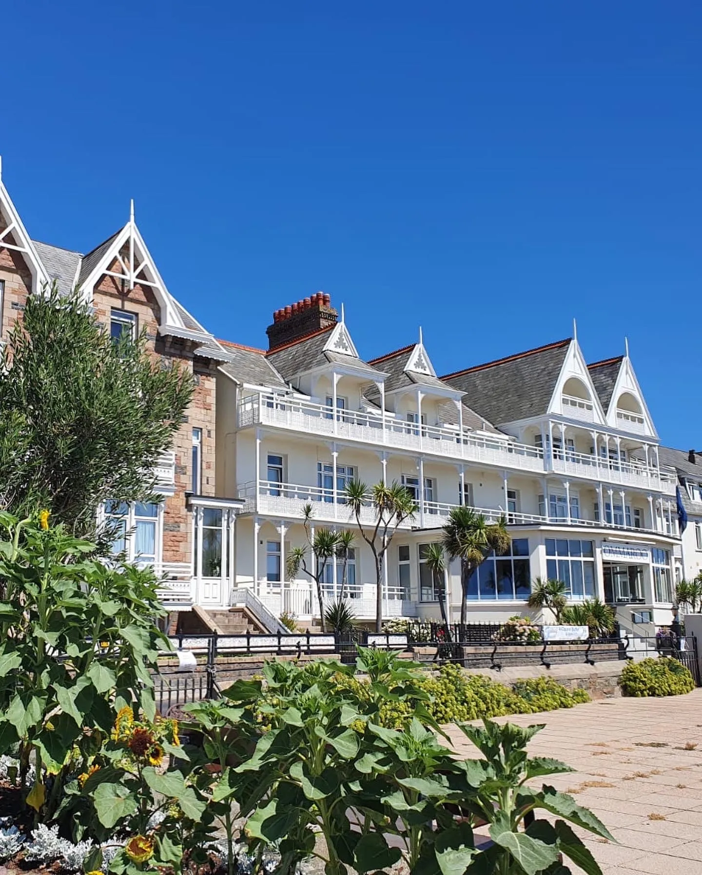 Exterior of Ommaroo Hotel with white balconies, decorative gables and seaside planting under a clear blue sky
