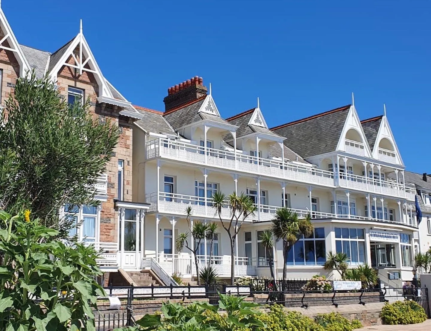 Exterior of Ommaroo Hotel with white balconies, decorative gables and seaside planting under a clear blue sky