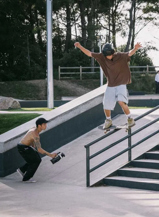 Skateboarder grinding down a handrail at an outdoor skate park while being filmed.