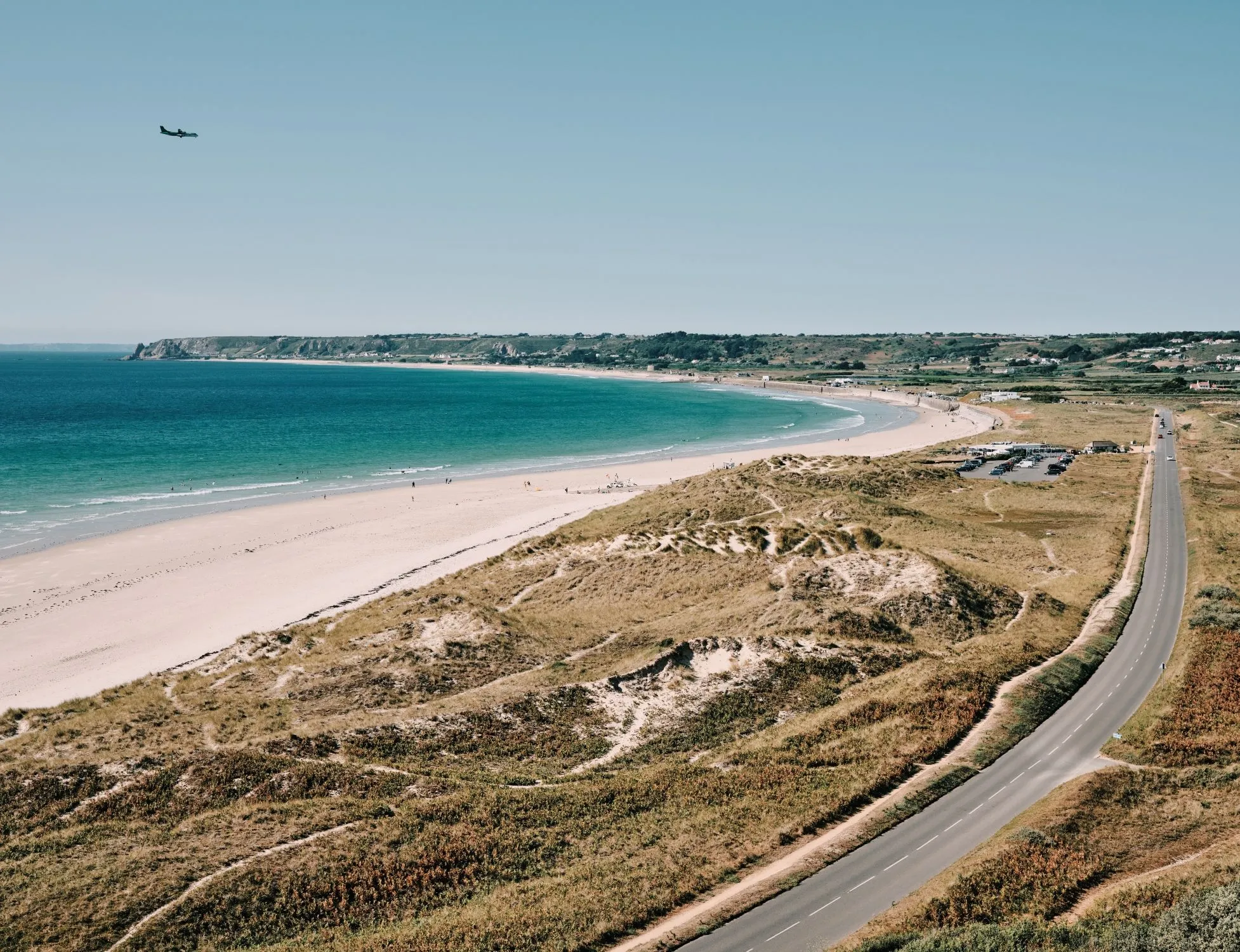 Aerial view of St Ouen's Bay with sandy beach, turquoise sea, sand dunes and a coastal road under a clear blue sky.