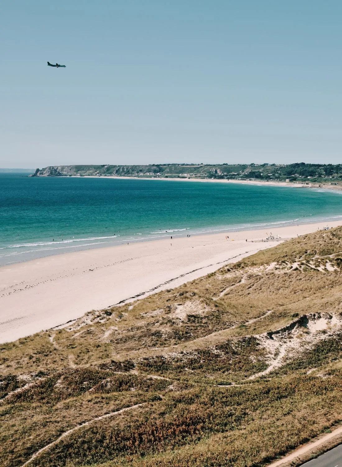 Aerial view of St Ouen's Bay with sandy beach, turquoise sea, sand dunes and a coastal road under a clear blue sky.