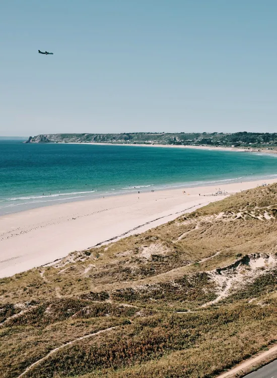 Aerial view of St Ouen's Bay with sandy beach, turquoise sea, sand dunes and a coastal road under a clear blue sky.