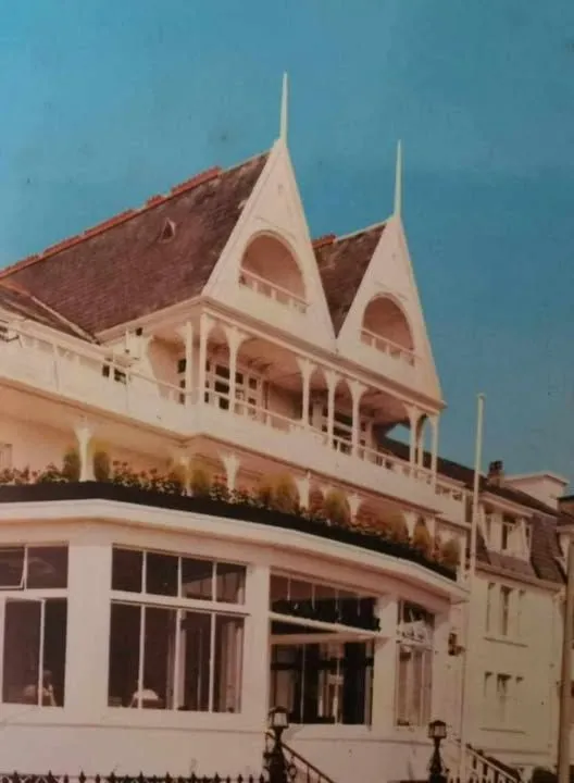 Historic photograph of a grand Victorian-style hotel with ornate balconies and pointed gables under a bright blue sky.