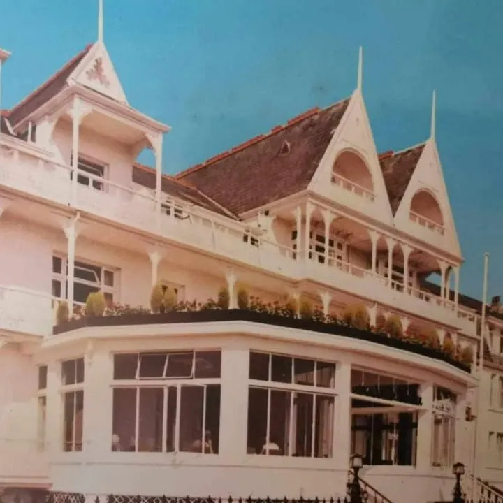 Historic photograph of a grand Victorian-style hotel with ornate balconies and pointed gables under a bright blue sky.