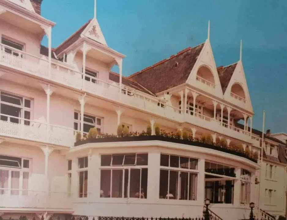 Historic photograph of a grand Victorian-style hotel with ornate balconies and pointed gables under a bright blue sky.