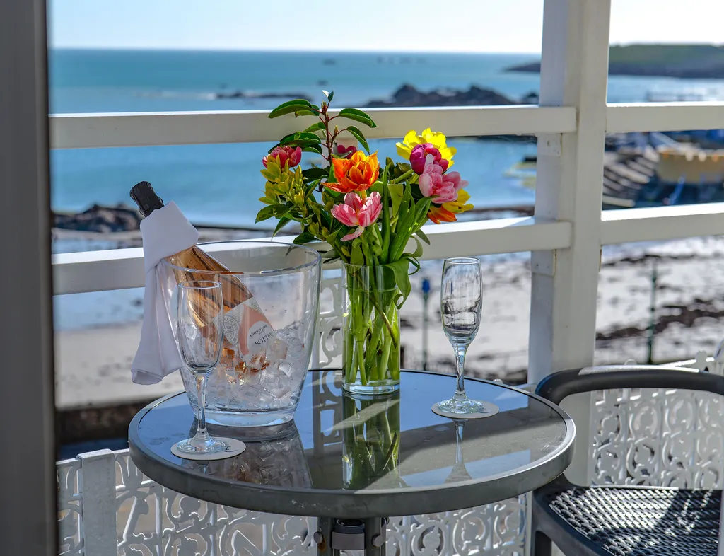 Ice bucket with a bottle of rosé, two flutes and a vase of colourful flowers on a balcony overlooking the sea.