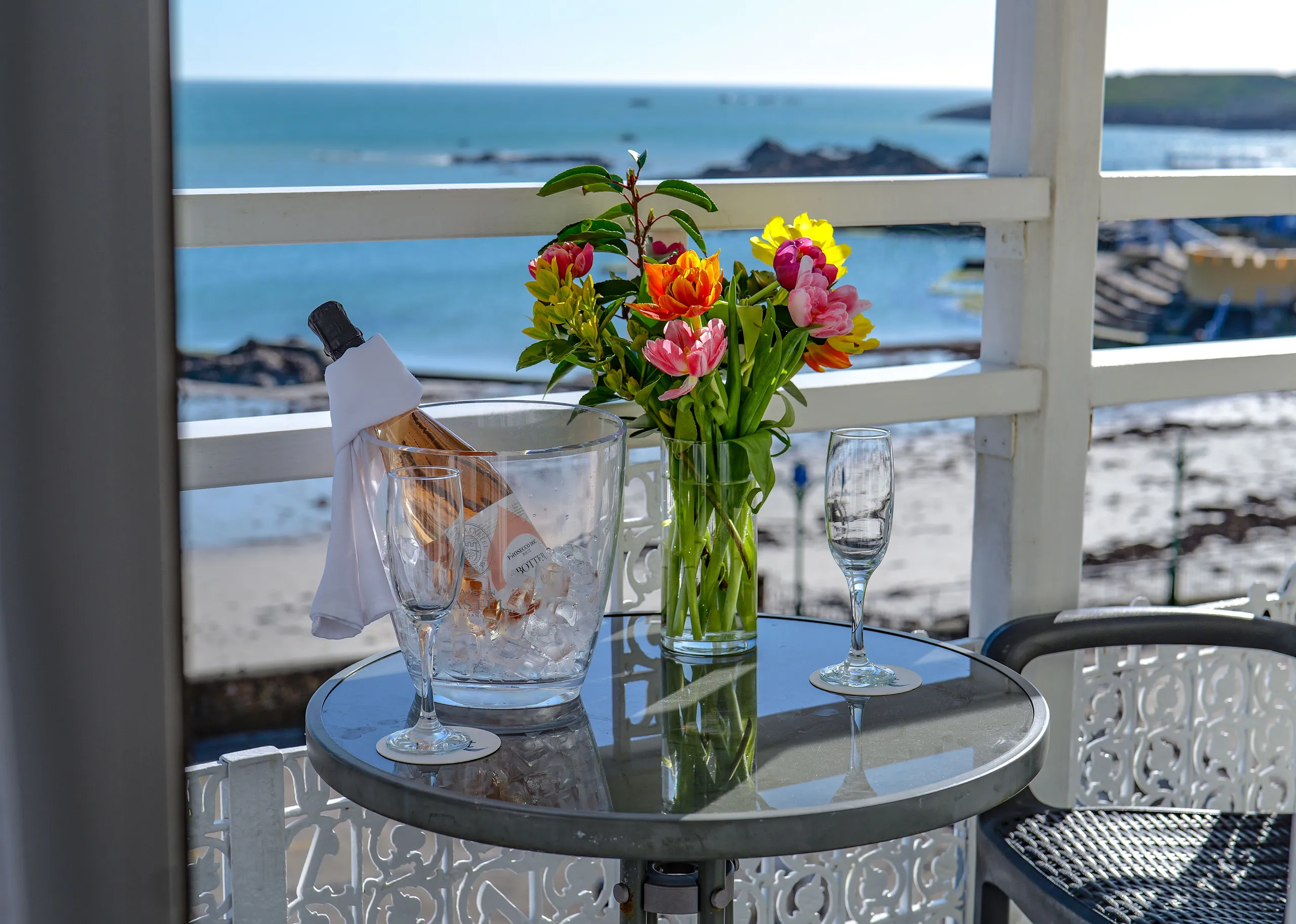 Ice bucket with a bottle of rosé, two flutes and a vase of colourful flowers on a balcony overlooking the sea.